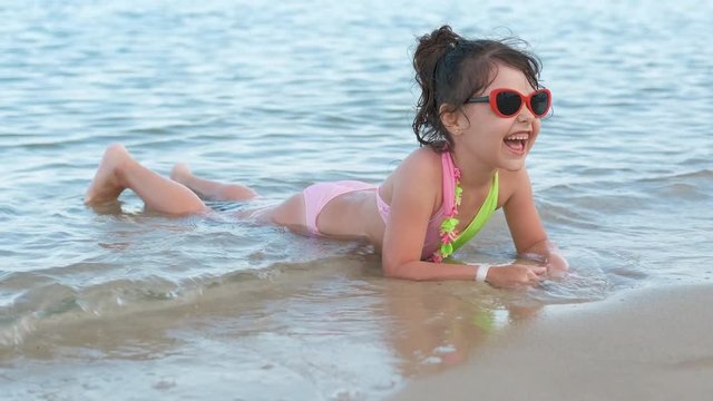 A happy child is lying at the seashore. Cheerful little girl in sunglasses lies on the seashore.