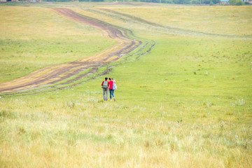 Fototapeta na wymiar Two young girls with backpacks and photo camera make selfie on phone near rural road. Horse farm pasture with mare and foal. Small village with old houses. Summer landscape with green hills