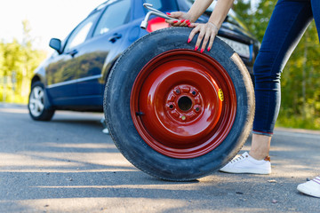 Closeup girl with spare wheel and cylinder key on the background of the car on the Jack. A woman with a spare wheel of a car with a flat wheel. Replacement tires on the side of the road.