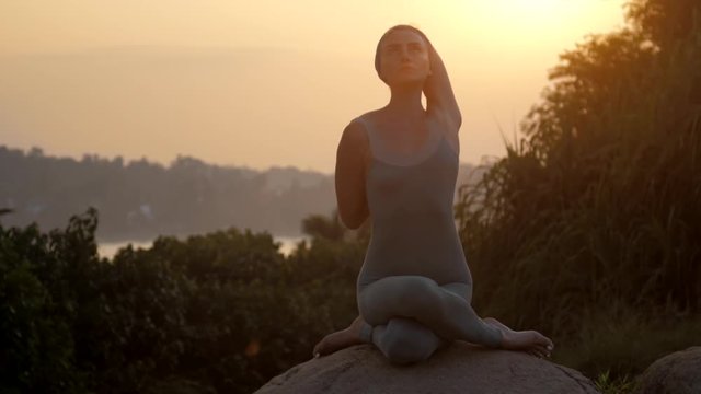 young yoga practitioner sits in gomukhasana position on huge old rock against red sunrise light low angle shot slow motion. Concept fitness yoga wellness lifestyle