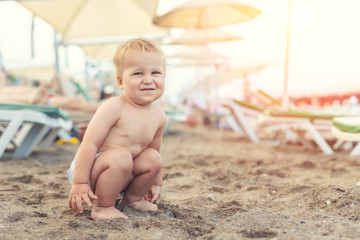 Cute caucasian toodler boy squatting alone on sandy beach between chaise-lounge. Adorable happy...