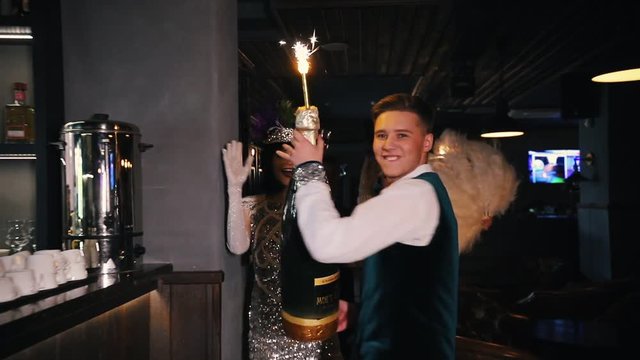 Retro theme party - A young man walking accompanied by two women in brilliant clothes - holding a huge bottle of champagne with fireworks on top