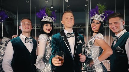 Theme party - People in glistening retro clothes are standing by their friend who raises the glass of drink up