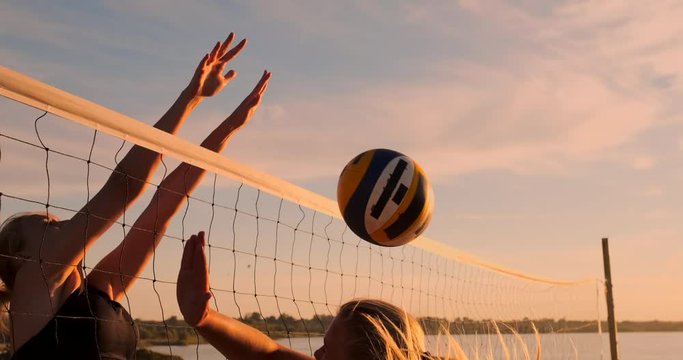 Young female volleyball players pass and spike the ball over the net on a sunny summer evening. Fit Caucasian girls playing beach volleyball at sunset