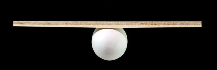 Balance concept, board on egg balance isolated on the black background, balancing on seesaw in...