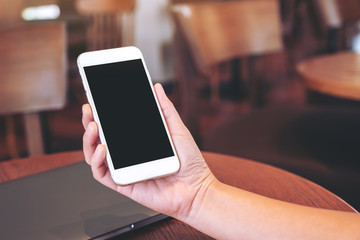 Mockup image of hands holding white mobile phone with blank desktop screen with laptop computer on the table