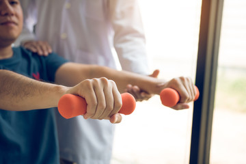 Asian physiotherapist helping a patient lifting dumbbells work through his recovery with weights in clinic room.