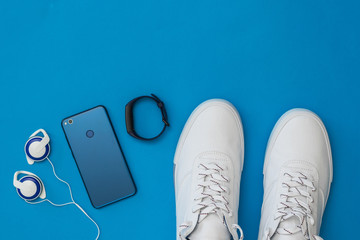 White sneakers, headphones, smart bracelet and blue smartphone on blue background. Sports style. Flat lay.