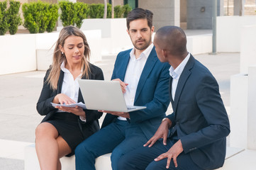 Business team checking slides on laptop before presentation. Mix raced group sitting outside, holding documents and open laptop and excited with discussion. Working together concept