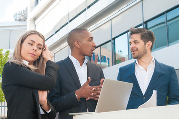 Creative team arguing while discussing startup project. Business people in office suits standing outdoors, using laptop and talking. Teamwork concept