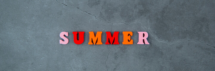 The multicolored summer word is made of wooden letters on a grey plastered wall background.