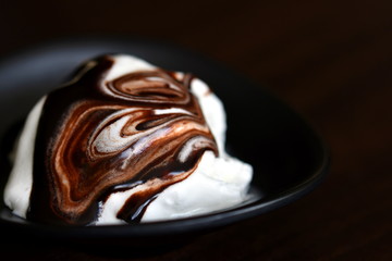 Dark brown melting chocolate dressing swirl on the white vanilla ice cream in dark bowl closeup....