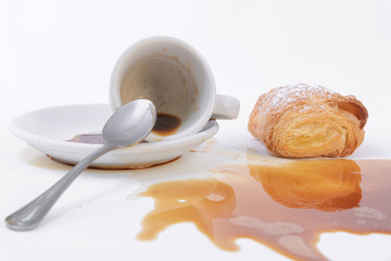 A tasty freshly baked golden puff pastry roll and a cup of coffee spilled over the table.