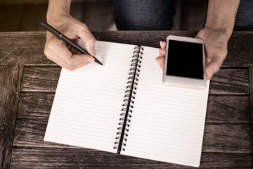 A right hand ofwomen holding a pen writing a notebook and left hands playing a smart phone. Recording and business concept.