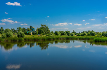 Blue sky with rare clouds reflected from the surface of the pond on the shore of which stands a blue Church surrounded by greenery