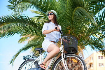 Close-up of attractive smiling girl in sunglasses, white shorts, blouse and hat riding a bicycle on green palm tree background on bright sunny day. Active lifestyle and vacations concept.