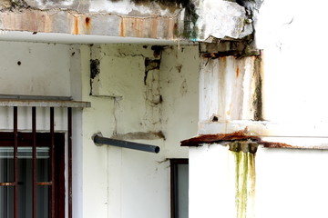 Dirty dilapidated cracked white corner building facade with rusted metal bars on windows and fallen concrete from above balcony caused by rain damage on rainy spring day