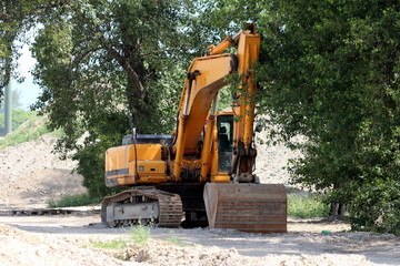 Fototapeta na wymiar Excavator parked in shade of large trees waiting for trucks to load at large construction site on warm sunny spring day