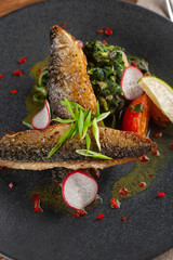 Close up of fried mackerel fish with vegetable garnish: spinach in coconut milk, radish, onion and baked tomatoes served on a moderm black plate