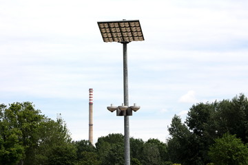 LED modern street light reflectors pointed towards large reflective panels mounted on top of strong metal pole with industrial chimney in background surrounded with dense trees on warm sunny spring da