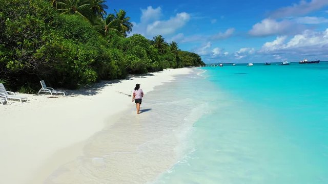 A woman is walking along  a beach through soft waves of the turquoise sea. A forest to the left. Boats and a blue cloudy sky in the distance. Dolly in truck right
