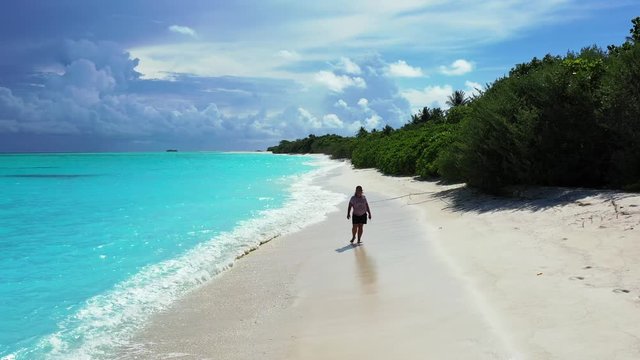A woman is walking along a beach through soft waves of the turquoise sea. A forest to the right. A cloudy sky in the distance. Dolly out