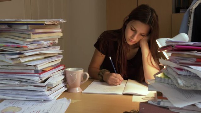 Static shot of a stressed lady sitting on a desk with a pile of books studying and doing her homework while writing on a notebook until she gets tired and sleeps