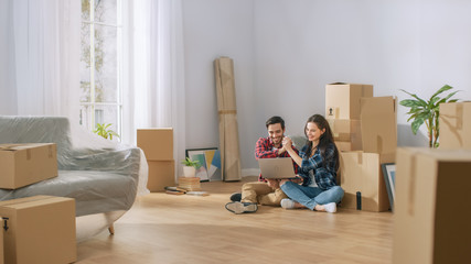 Happy Young Couple Sitting on Floor of Newly Rented / Purchased Apartment Use Laptop Computer. Successful Boyfriend and Girlfriend do High Five. Cardboard Boxes and Covered Furniture in Bright Home