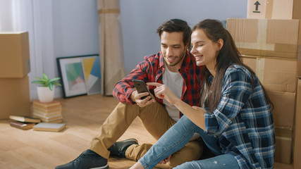 Happy Young Couple Sitting on the Floor of the Newly Rented / Purchased Apartment Use Smartphone. Unpacked Cardboard Boxes and Covered Furniture in the Modern Bright Sweet Home.