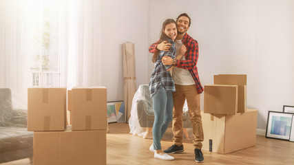 Young Couple Moving Into New Apartment, Carrying Cardboard Boxes with Stuff, Having Fun and Hugging. Boyfriend and Girlfriend Start Living Together, Unpacking Stuff. Room Lit by Warm Yellow Sunlight