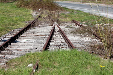 Overgrown old unused rusted railroad tracks resting on wooden railroad ties next to paved road on warm sunny spring day