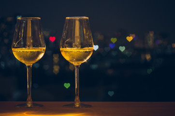 Two glasses of white wine on wooden table of rooftop bar with colorful love shape bokeh of city light and space for text.
