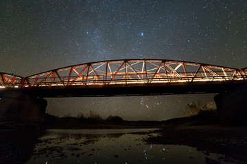 Illuminated metal bridge on concrete supports reflected in water on dark starry sky with Milky Way constellation background. Night photography concept.