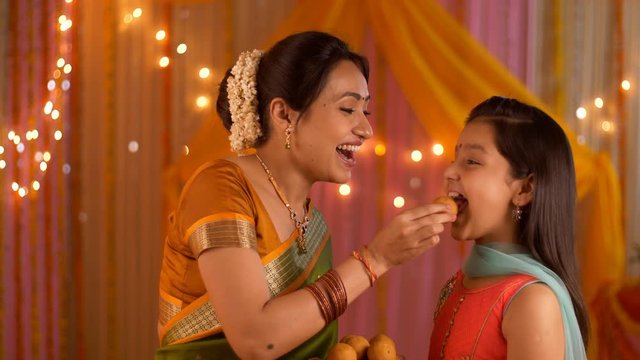 Smiling Indian mother feeding sweets to her young daughter during festivities - Happy Family. Indian stock video of a mother wishing daughter on the auspicious day of Diwali festival / Durga Puja 