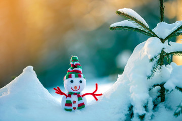 Small funny toy baby snowman in knitted hat and scarf in deep snow outdoors on bright blue and...