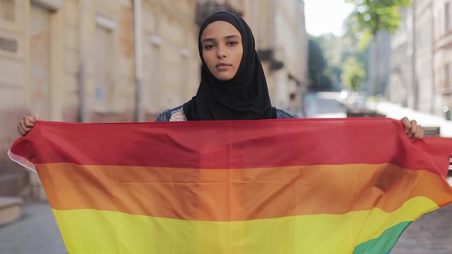 Portrait of beautiful young Muslim woman wearing hijab headscarf with LGBT flag standing on the old street background. Lesbianism and Islam concept.