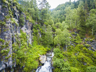 Fototapeta na wymiar A large waterfall in the back of the Altai Mountains with gray-brown stones near a steep cliff with green trees. Waterfalls Korbu aerial view.