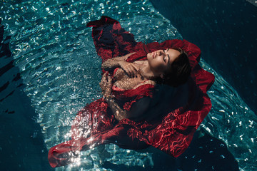Fashion photo: Girl  with bright make up in a red dress lying on the water of the pool. Young woman...