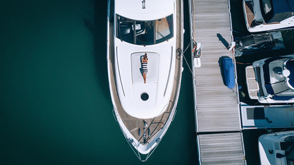 Aerial photo of a Young lady in a striped dress relaxing on board of the yacht near pier.