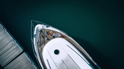 Aerial photo of a tropical woman in a striped dress relaxing on nose of the yacht near pier.