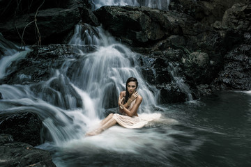 Fototapeta na wymiar Sensual female in white dress is sitting in the water on a big waterfal