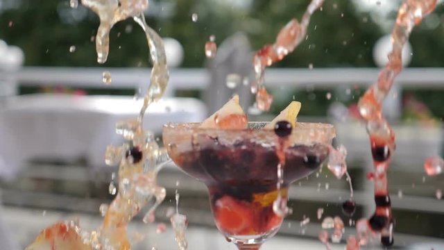 Fresh summer cocktail concept. Berries and ice cubes splash falling down into gluss with beautifully decorated fruit cocktail , shot in slow motion on blurred restaurant open summer terrace. Food