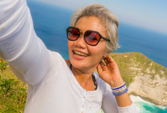 middle age 50s happy and cheerful Asian woman with grey hair taking selfie with mobile phone at beautiful tropical beach island smiling at cliff viewpoint enjoying Summer