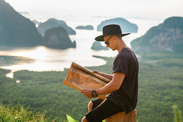 Handsome Young Man Looking at Paper Map Outdoors. Casually dressed Man Holding Map. Beautiful view of Nature. Young Traveler Enjoying Sunny Day. Traveling, Vacation and Freedom Concept -