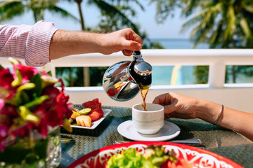 Breakfast for a young couple at a tropical resort close-up. A man pours tea to a woman. On the table is a plate with tropical fruits. Summer holidays in hot countries