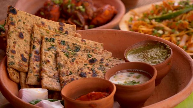 Paratha served with pickle, green dip, curd chutney, onion pieces and a chilli. The clay plate in the video is rotating.