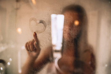 Beautiful woman taking a shower and drawing heart on the sweat and blurred glass.