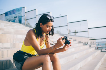 Young Spanish hipster girl in casual wear checking pictures on photo technology while resting on...