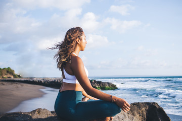 Fototapeta na wymiar Dark skinned woman calms brain minds with lotus yoga pose enjoying morning training on beach coastline, African American female increases awareness and attentiveness with padmasana exercise