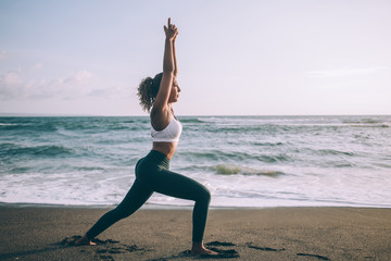 Fototapeta na wymiar Attractive young woman in casual tracksuit doing virabhadrasana one exercise during outdoors training near sea, female enjoying yoga healthy lifestyle for relaxing minds and get soul harmony
